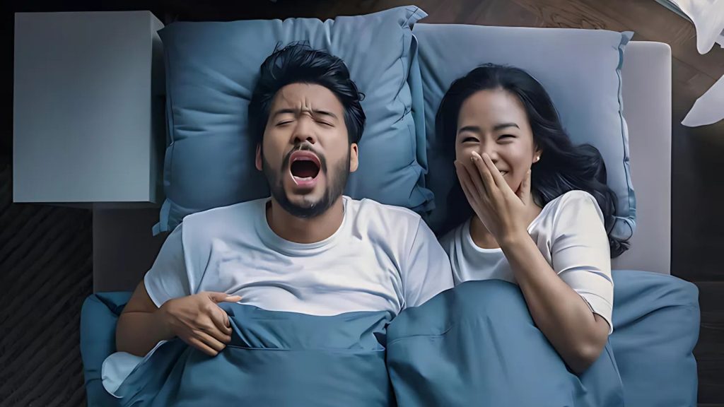 Man yawning loudly while woman laughs beside him in bed.